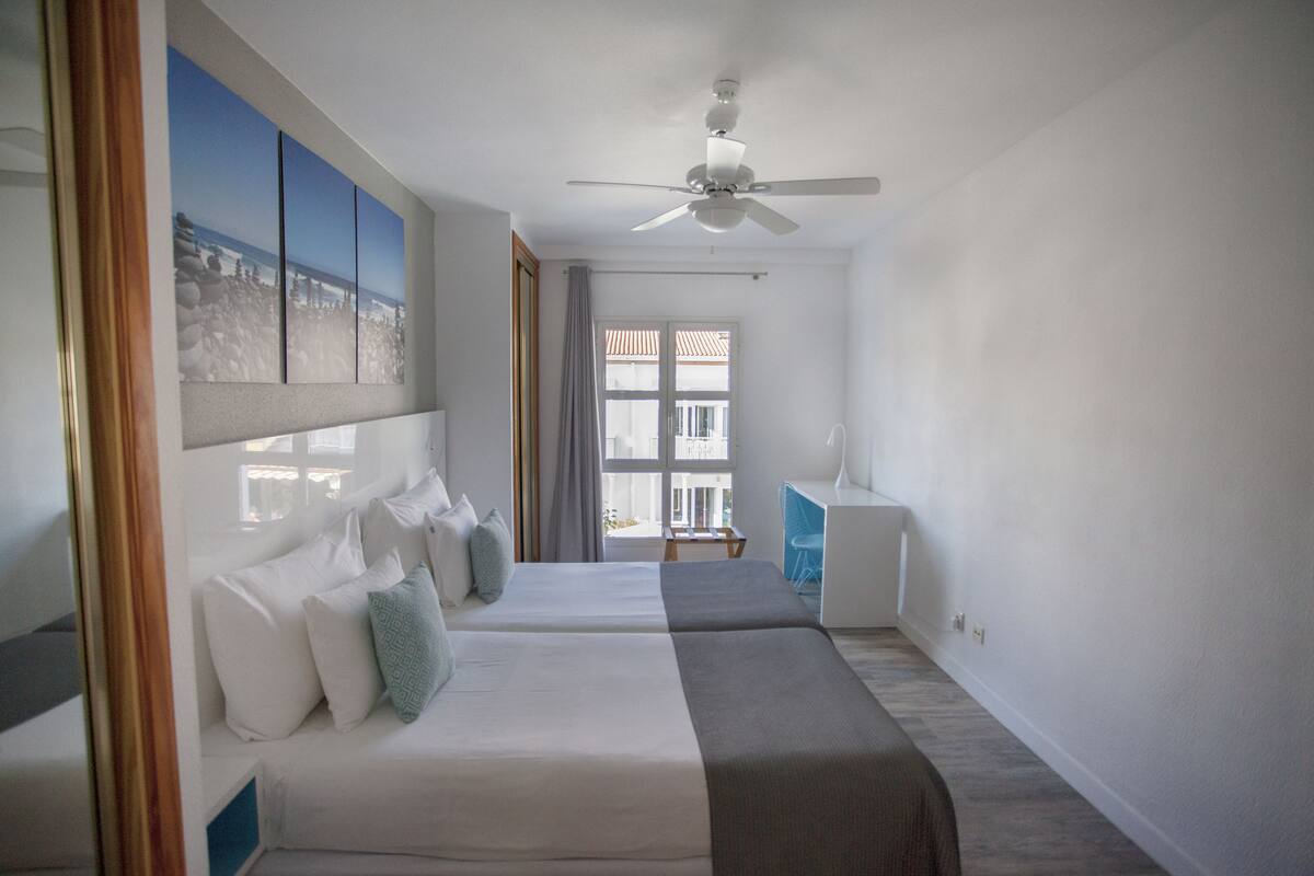 Habitación de hotel con cama, escritorio, silla, ventilador de techo y ventana con cortinas.