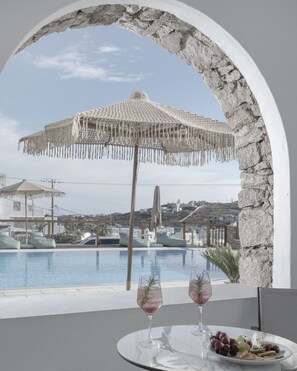 A poolside view with a stone archway, a woven umbrella, and a table with a glass of wine and a fruit plate. - Mykonos Essence - Adults Only (Mykonos)