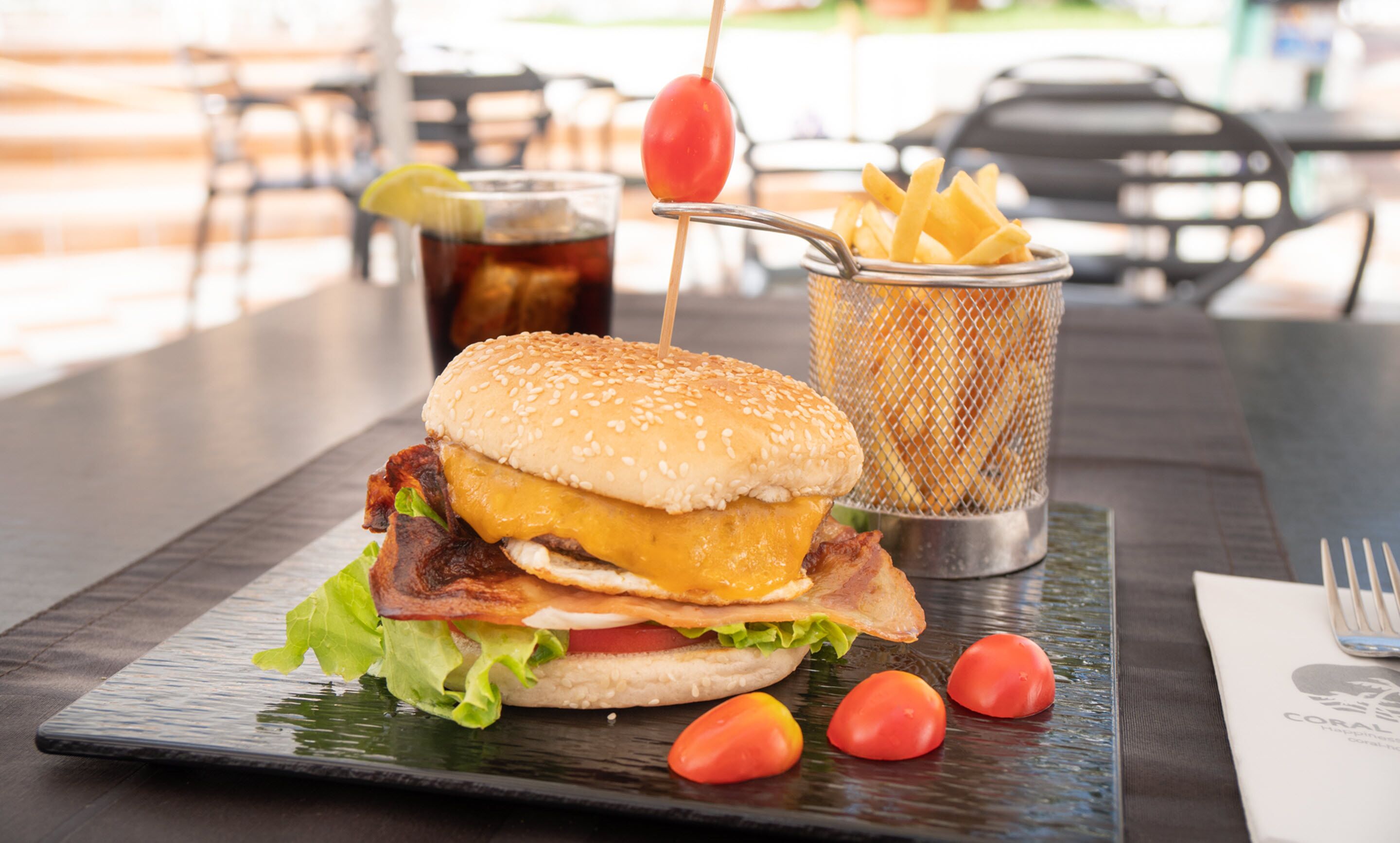 A burger with bacon, cheese, and lettuce on a plate, accompanied by fries and a drink.