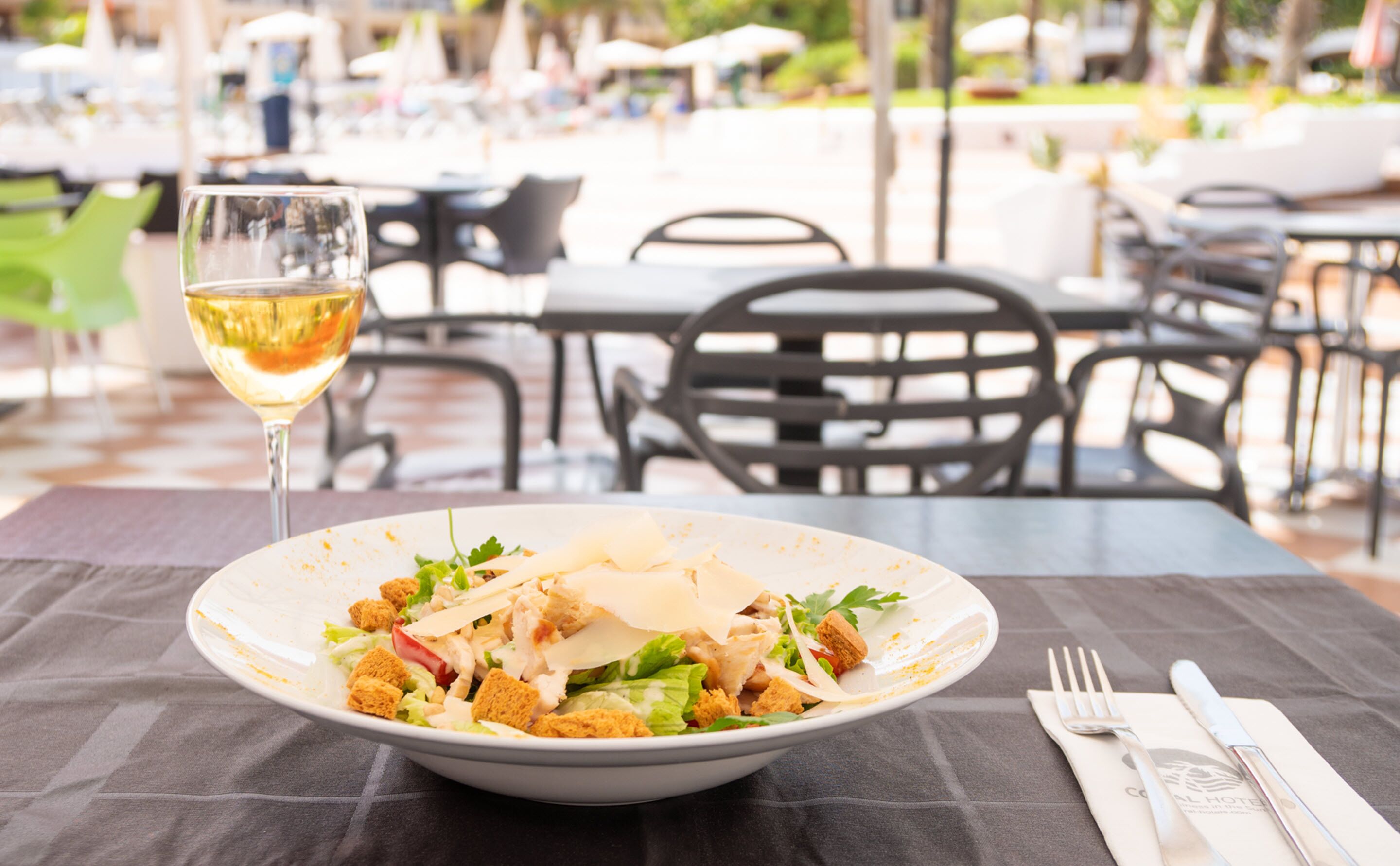 A dining table with a bowl of salad, a glass of white wine, and a fork and knife on a tablecloth.