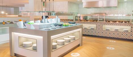 A modern buffet setup with a central island, multiple serving stations, and a kitchen area in the background.