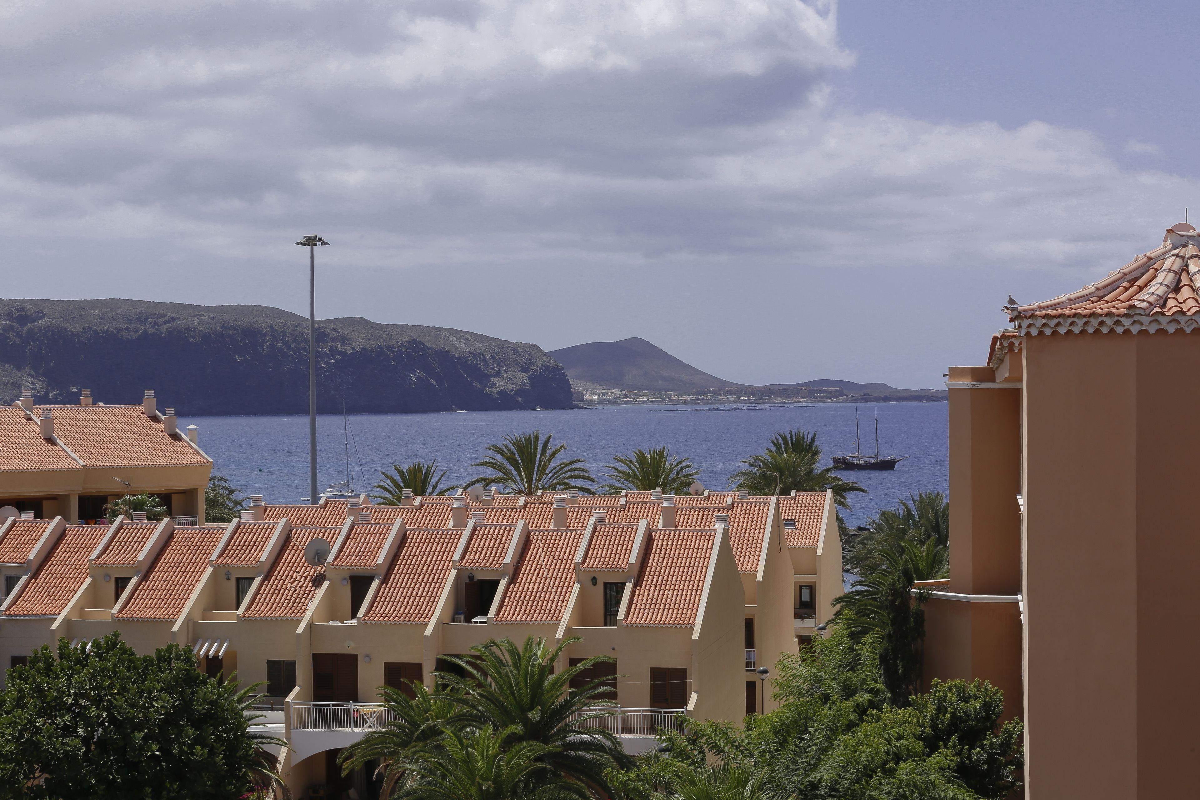 A coastal hotel with terracotta tiled roofs, a view of the sea, and mountains in the background.