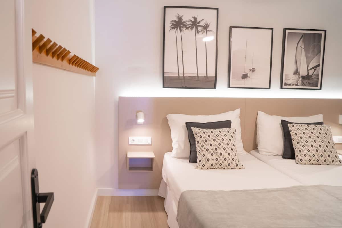 A hotel room with a bed, bedside tables, a nightstand, a wall-mounted light, and framed artwork.