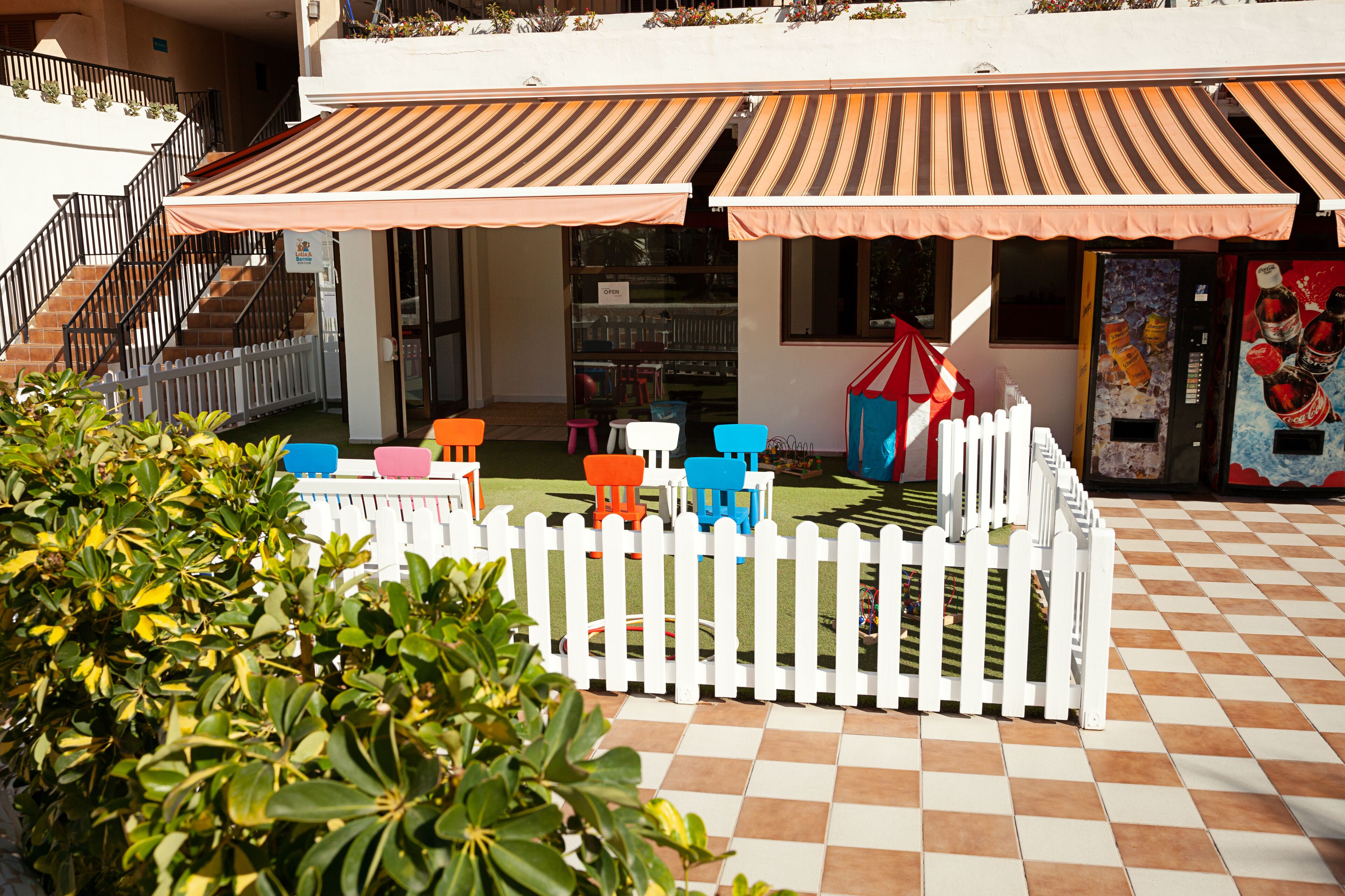 A courtyard with a white picket fence, outdoor seating area with colorful chairs, and a vending machine.