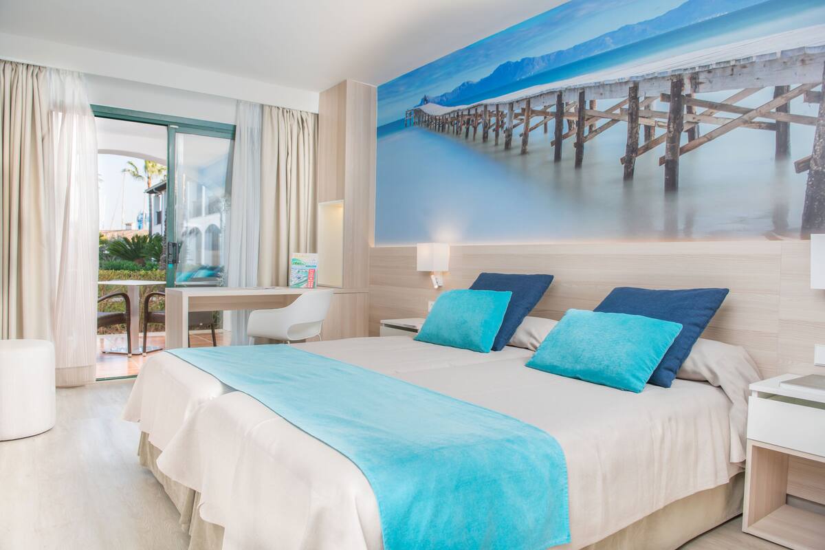 A hotel room with a large bed, a desk, and a view of a pier through a glass door.
