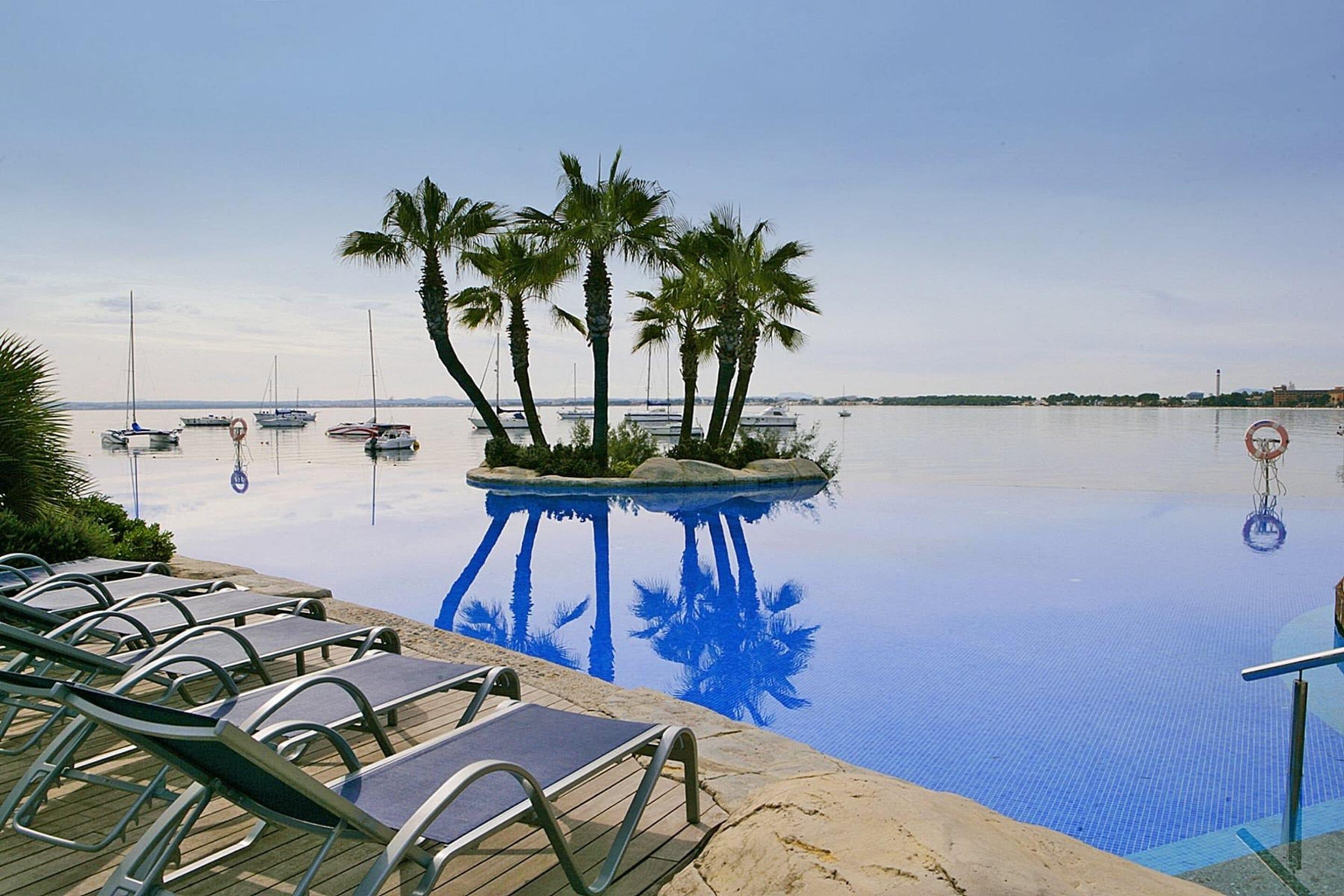Una piscina con sillas de descanso, palmeras y barcos al fondo.