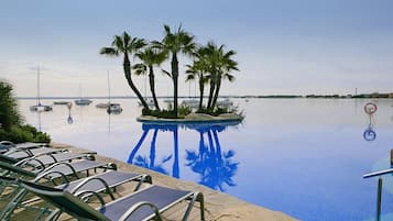 Una piscina con sillas de descanso, palmeras y barcos al fondo.
