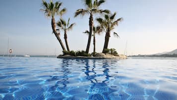 Ein Swimmingpool mit klarem, blauem Wasser, umgeben von Palmen und im Hintergrund eine Ansicht auf Boote und eine Brücke.