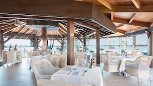 A restaurant with wicker furniture, white tablecloths, and a view of boats on the water. - Alcudiamar Marina Residence (Alcúdia)