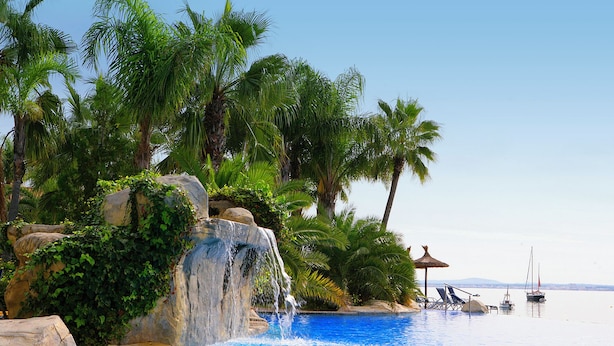 A pool with a waterfall, surrounded by palm trees and a clear sky. - Alcudiamar Marina Residence (Alcúdia)