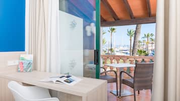Habitación de hotel con escritorio, una silla, un vidrio que da a un balcón con vista a palmeras y barcos.