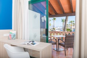 A hotel room with a desk, a chair, a glass door leading to a balcony with a view of palm trees and boats. - Alcudiamar Marina Residence (Alcúdia)