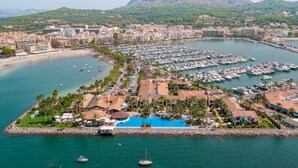 An aerial view of a marina with numerous boats docked, a beach area with palm trees, and a mountainous backdrop. - Alcudiamar Marina Residence (Alcúdia)