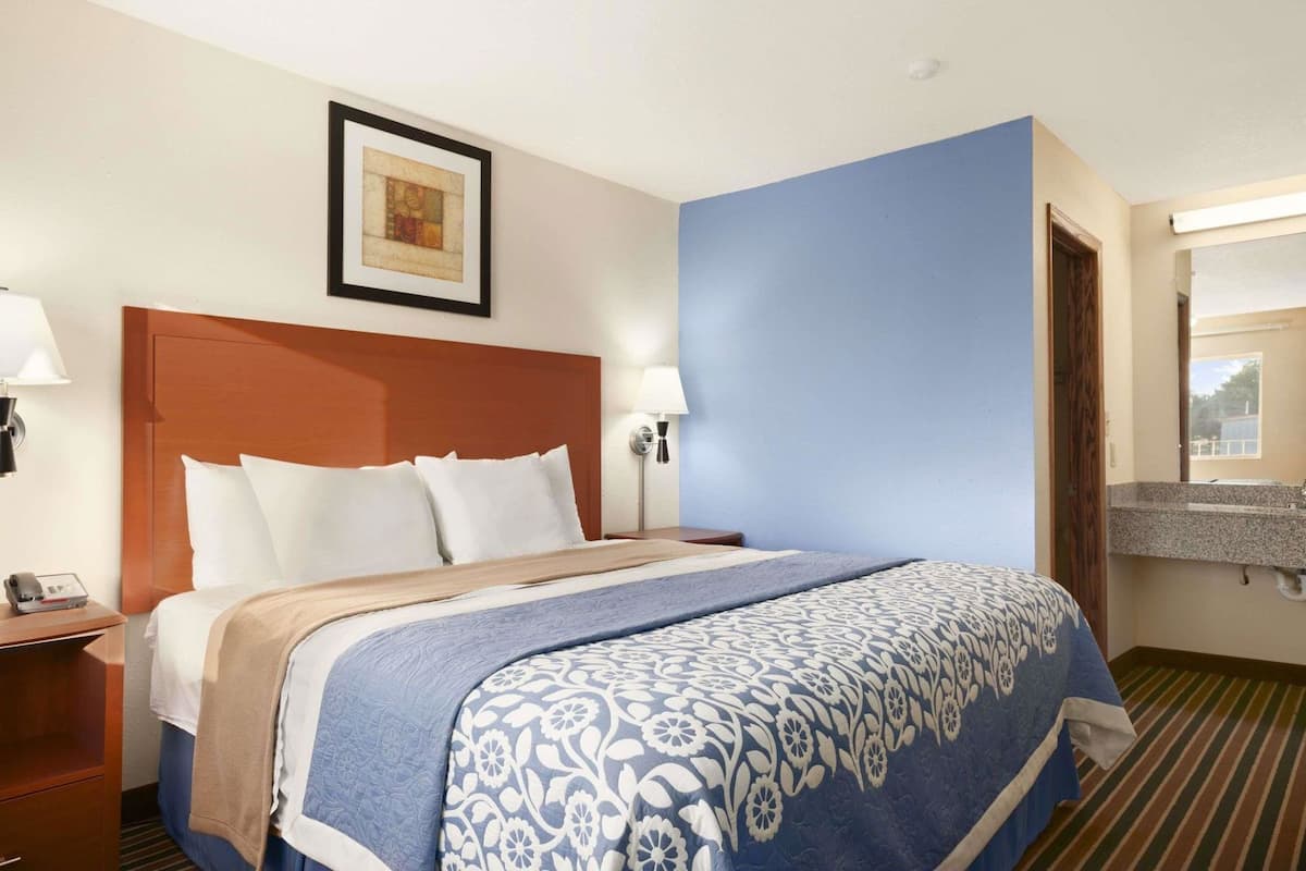 Ein Hotelzimmer mit Bett, Nachttischen, einem gerahmten Bild an der Wand und einem Badezimmer mit Spiegel und Waschbecken.