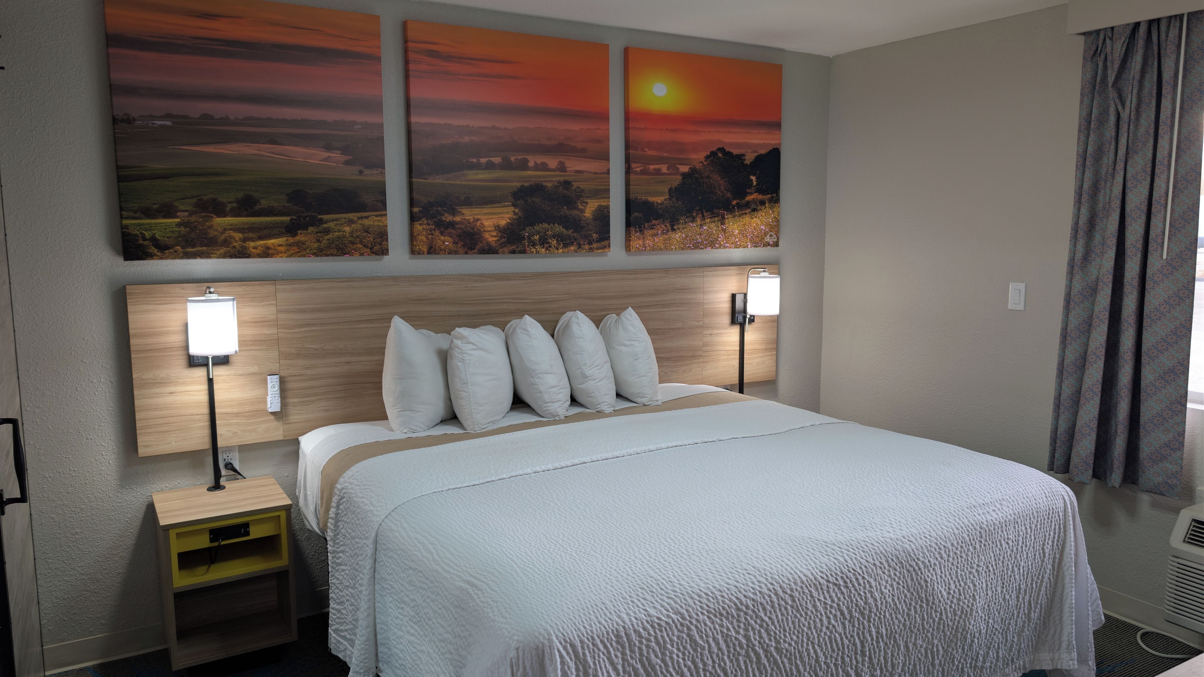 A hotel room with a large bed, two bedside lamps, a nightstand, and a wall-mounted artwork depicting a sunset.
