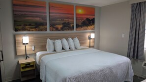 A hotel room with a large bed, two bedside lamps, a nightstand, and a wall-mounted artwork depicting a sunset. - Days Inn by Wyndham Champaign/Urbana (Champaign)