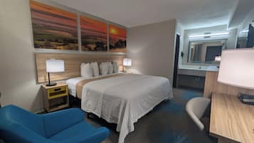 A hotel room with a bed, a desk, a chair, a nightstand, a lamp, a mirror, and a wall with a sunset painting.