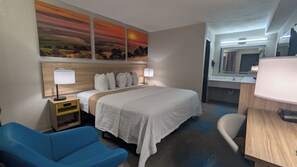 A hotel room with a bed, a desk, a chair, a nightstand, a lamp, a mirror, and a wall with a sunset painting. - Days Inn by Wyndham Champaign/Urbana (Champaign)