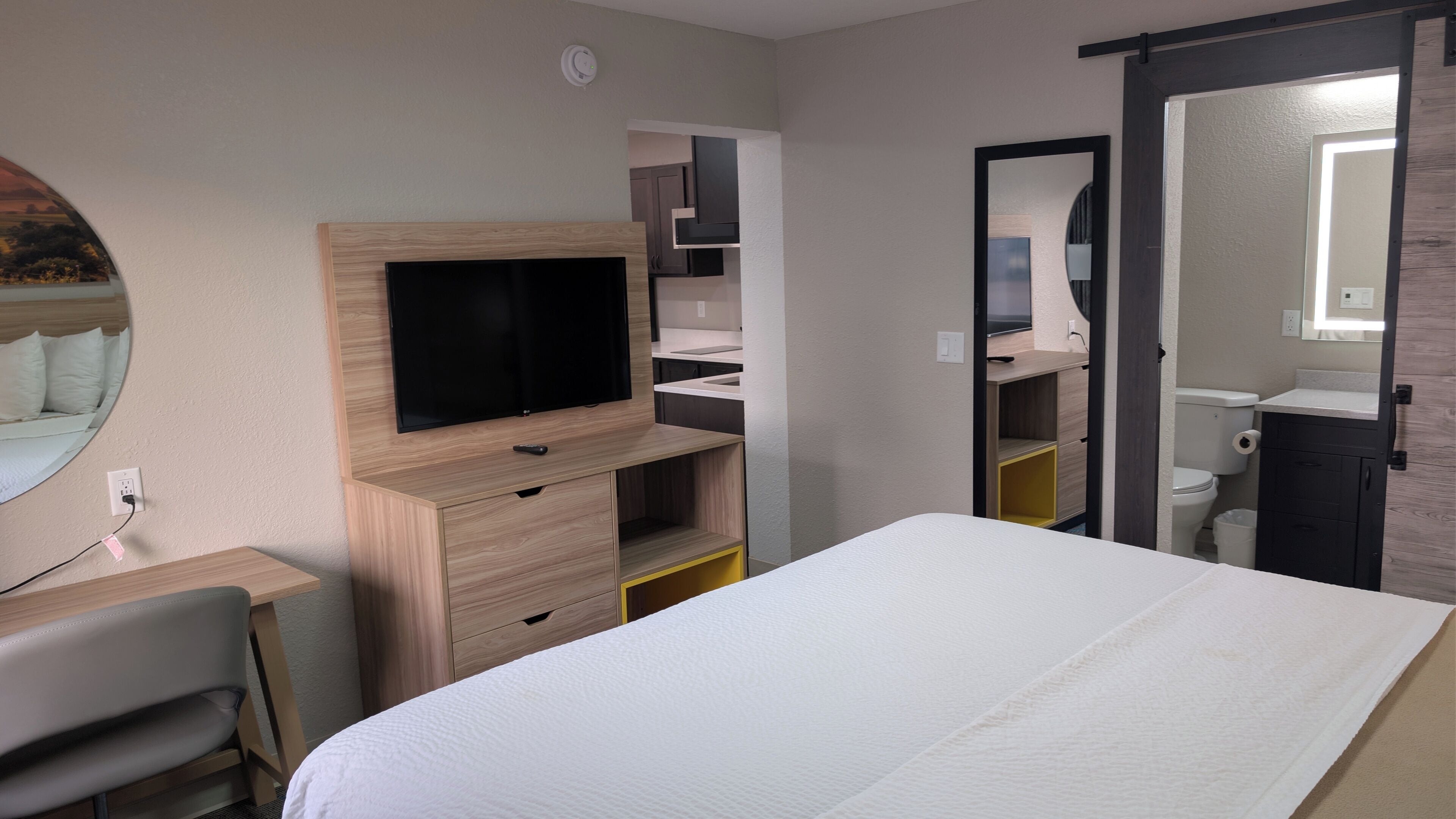 Habitación de hotel con cama, televisor montado en una cómoda de madera, escritorio con silla, espejo y baño visible a través de una puerta abierta.
