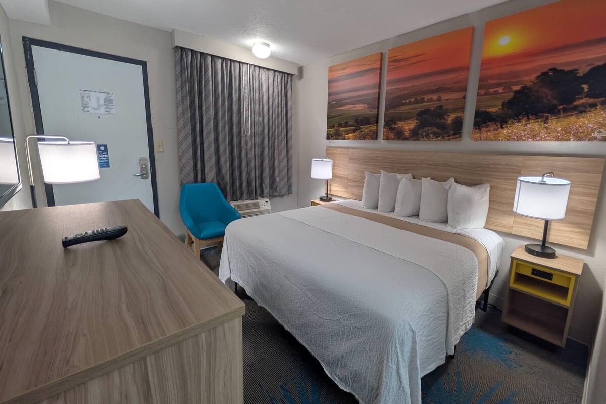 Ein Hotelzimmer mit Bett, Schreibtisch, Stuhl, Nachttisch und einer Wand mit Sonnenuntergangsbildern.