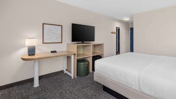 Hotellihuone, jossa on sänky, kirjoituspöytä lampun kanssa, televisio ja roskakori.