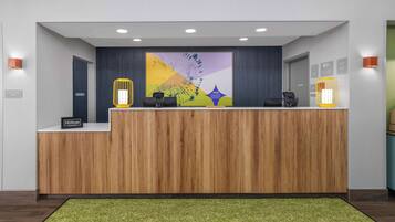 Un mostrador de recepción de hotel con detalles de madera, un cuadro abstracto de gran tamaño encima y una alfombra verde al frente.