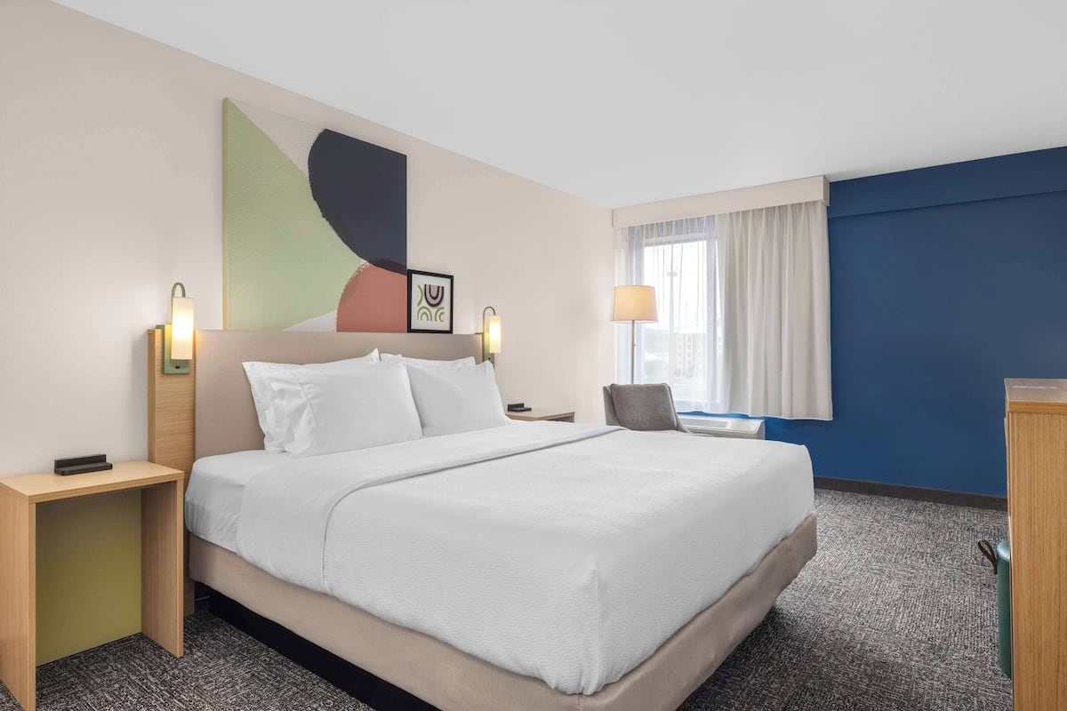 A hotel room with a large bed, two bedside tables, a desk, a chair, a window with curtains, and wall art.