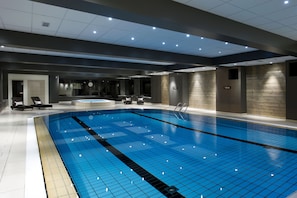A modern indoor swimming pool with a blue tiled floor, lounge chairs, and a staircase leading to upper levels.