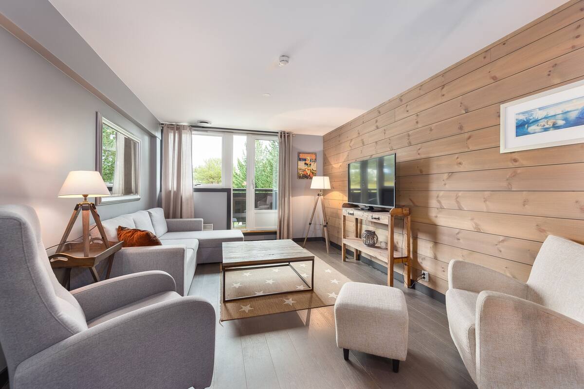A modern living room with grey sofas, a wooden coffee table, and a TV on a wooden wall.