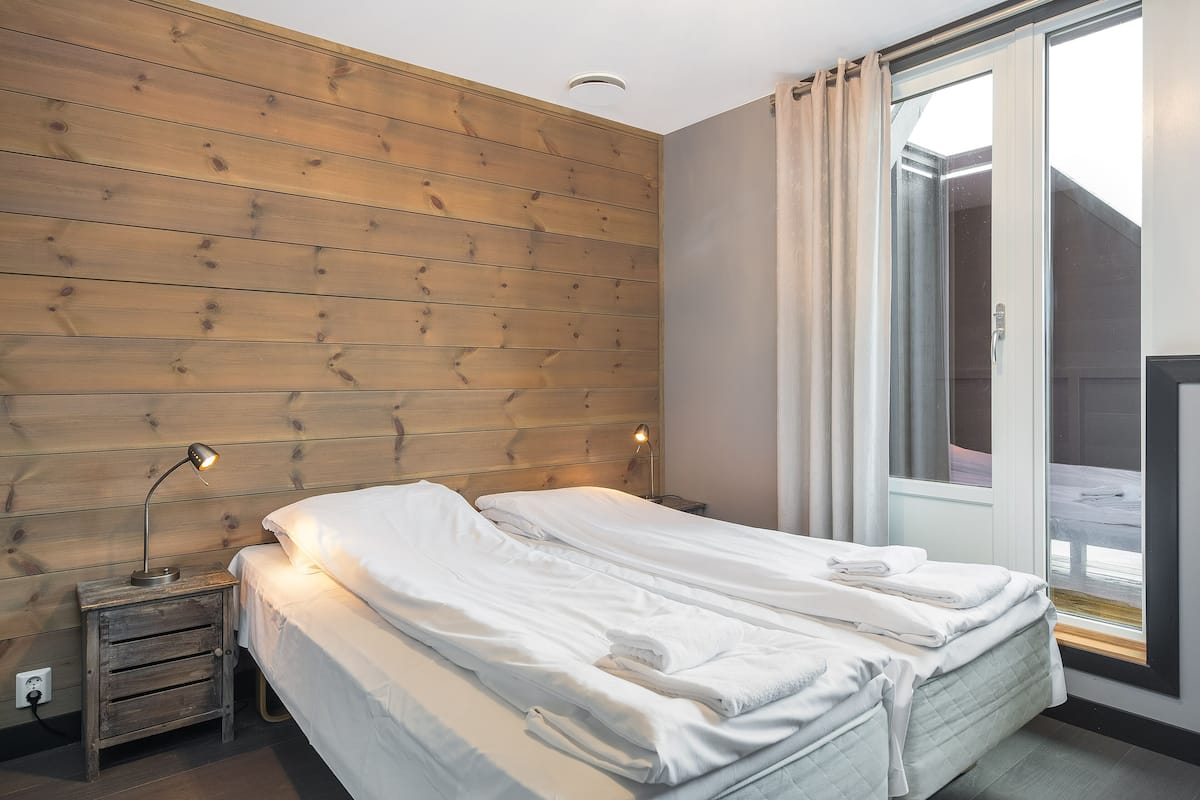 A bedroom with a wooden headboard, a bed with white linens, a bedside table with a lamp, and a window with curtains.