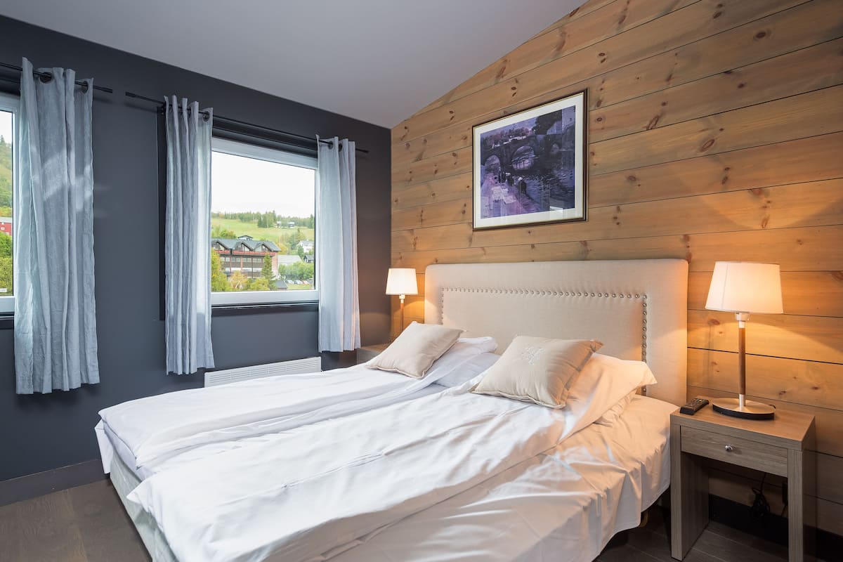 A bedroom with a large bed, two bedside lamps, a nightstand, and a window with curtains.