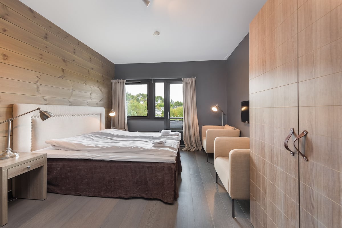 A modern bedroom with a wooden headboard, a large bed, bedside lamps, a seating area, and a view of greenery outside.