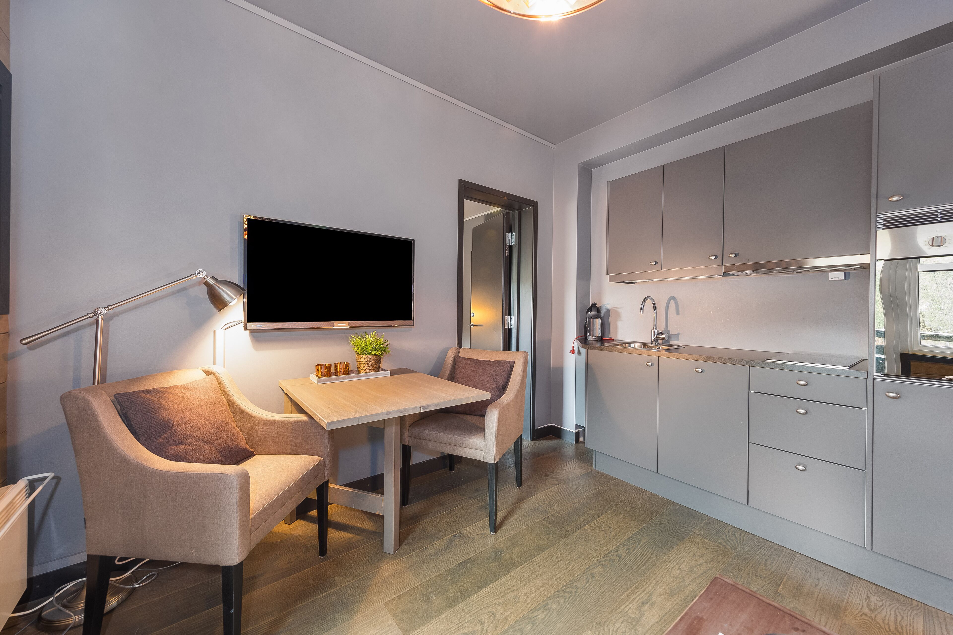 A modern kitchen with grey cabinets, a wooden dining table, and a flat-screen TV mounted on the wall.