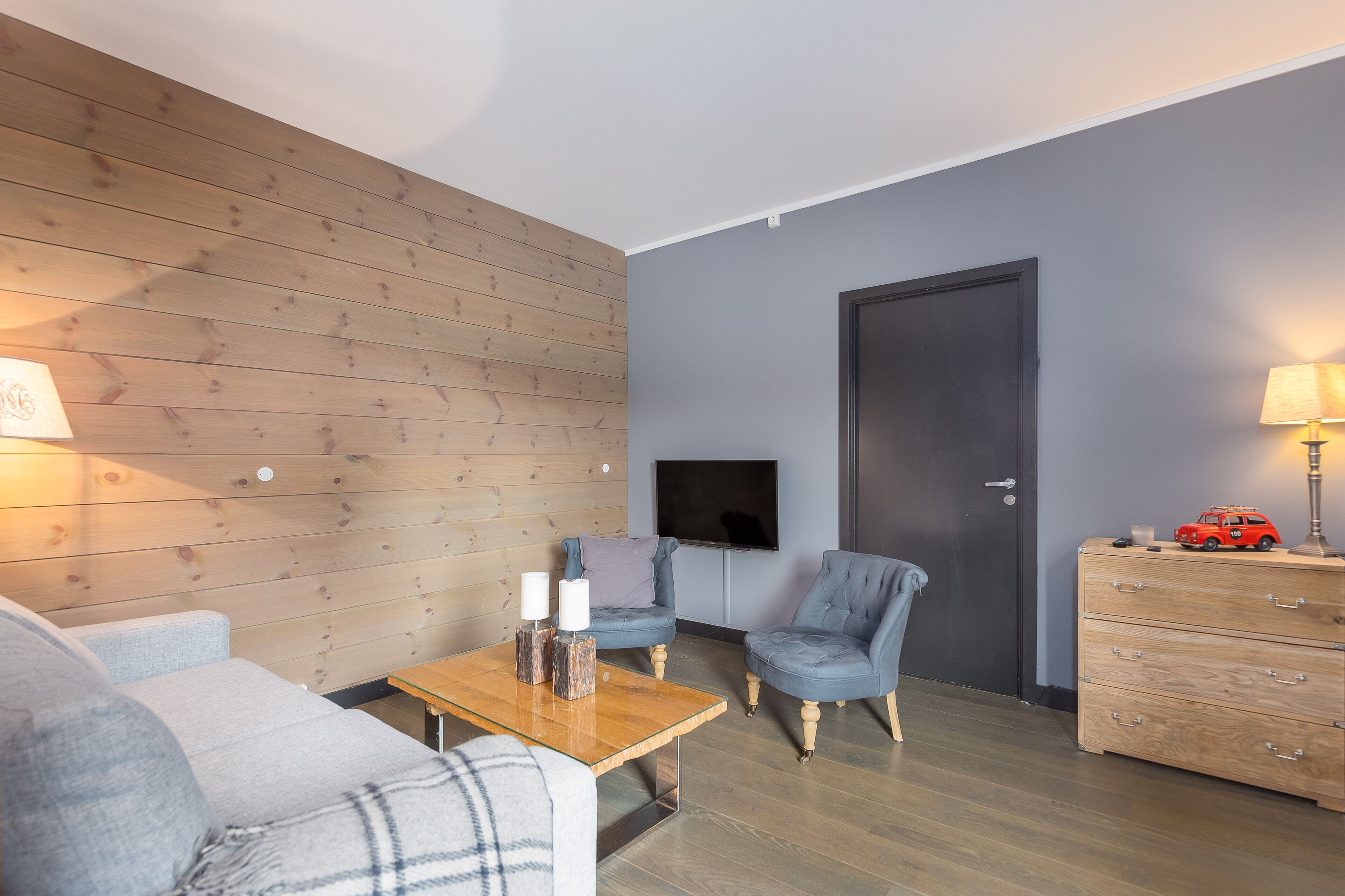 Una sala de estar moderna con una pared de madera, un sofá gris, un sillón azul, una mesa de centro de madera y un televisor montado en la pared.