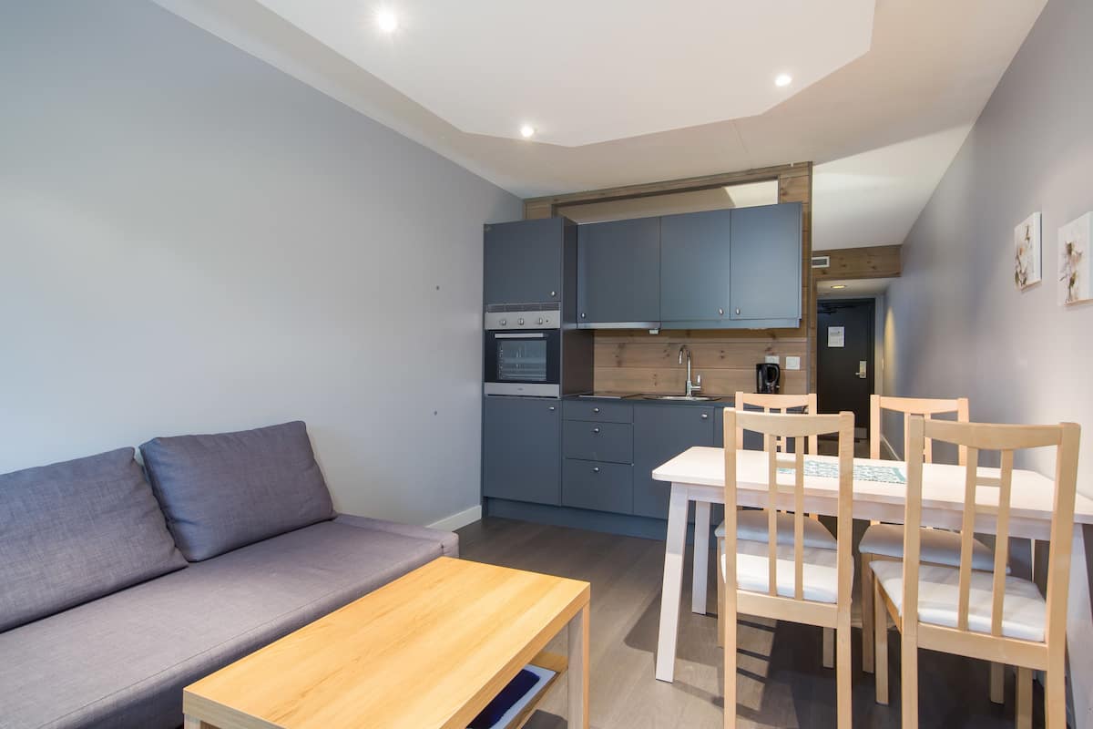 A compact living space with a sofa, a wooden table, a kitchen area with cabinets and appliances, and a dining table with chairs.
