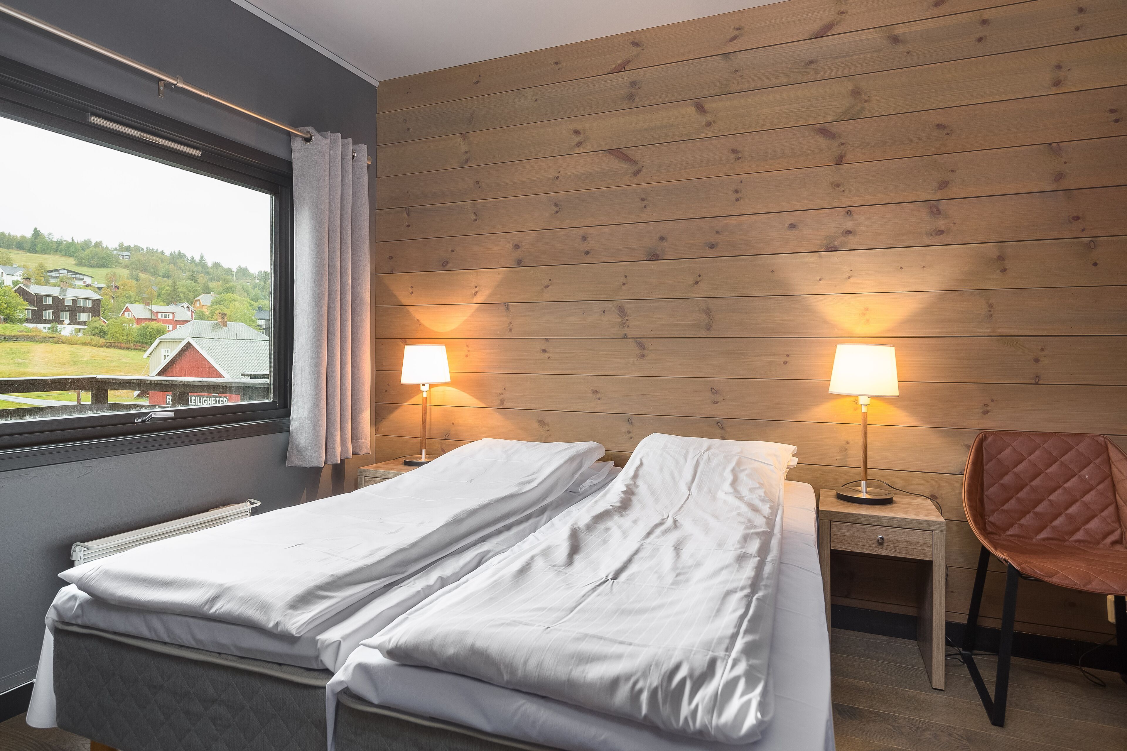 A bedroom with a bed, two bedside tables with lamps, a chair, and a window with a view of houses and greenery.