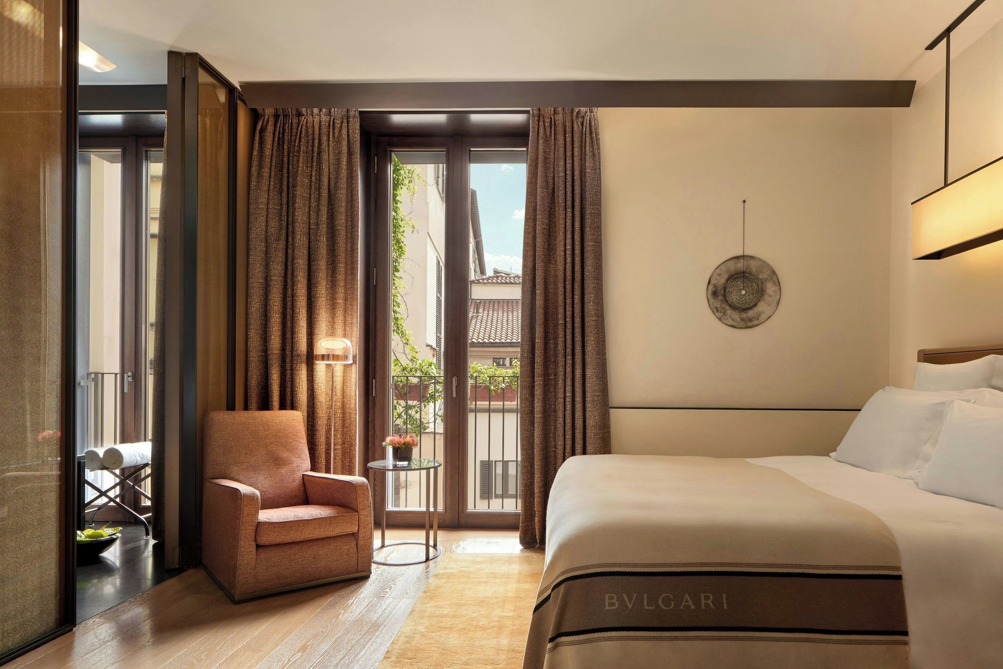 Habitación de hotel con una cama grande, un área de estar con una silla y una mesita, y vistas al exterior por una puerta corredera.