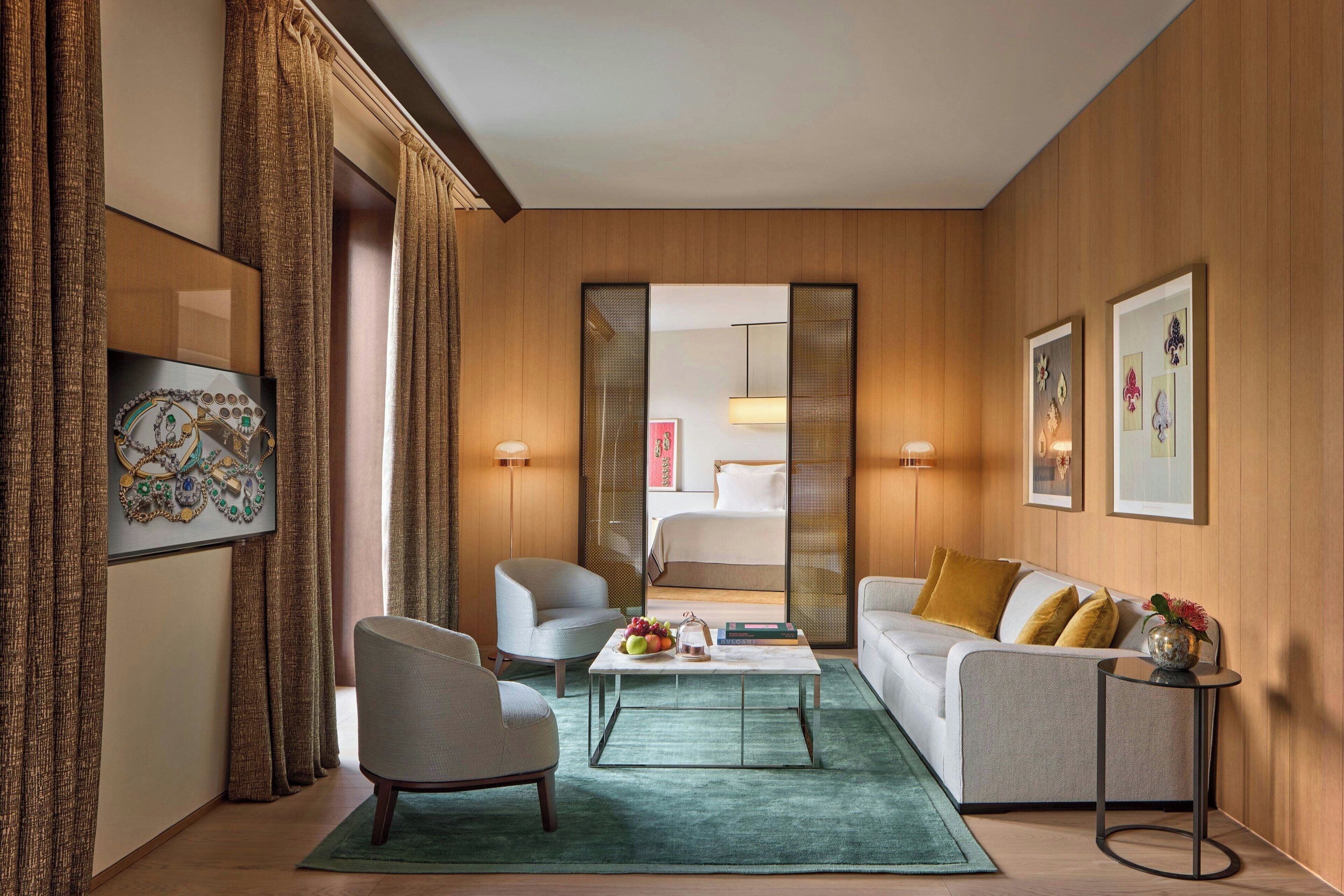 A modern hotel room with a sofa, armchair, and a coffee table. There is a bed visible through a glass door, and artwork on the walls.