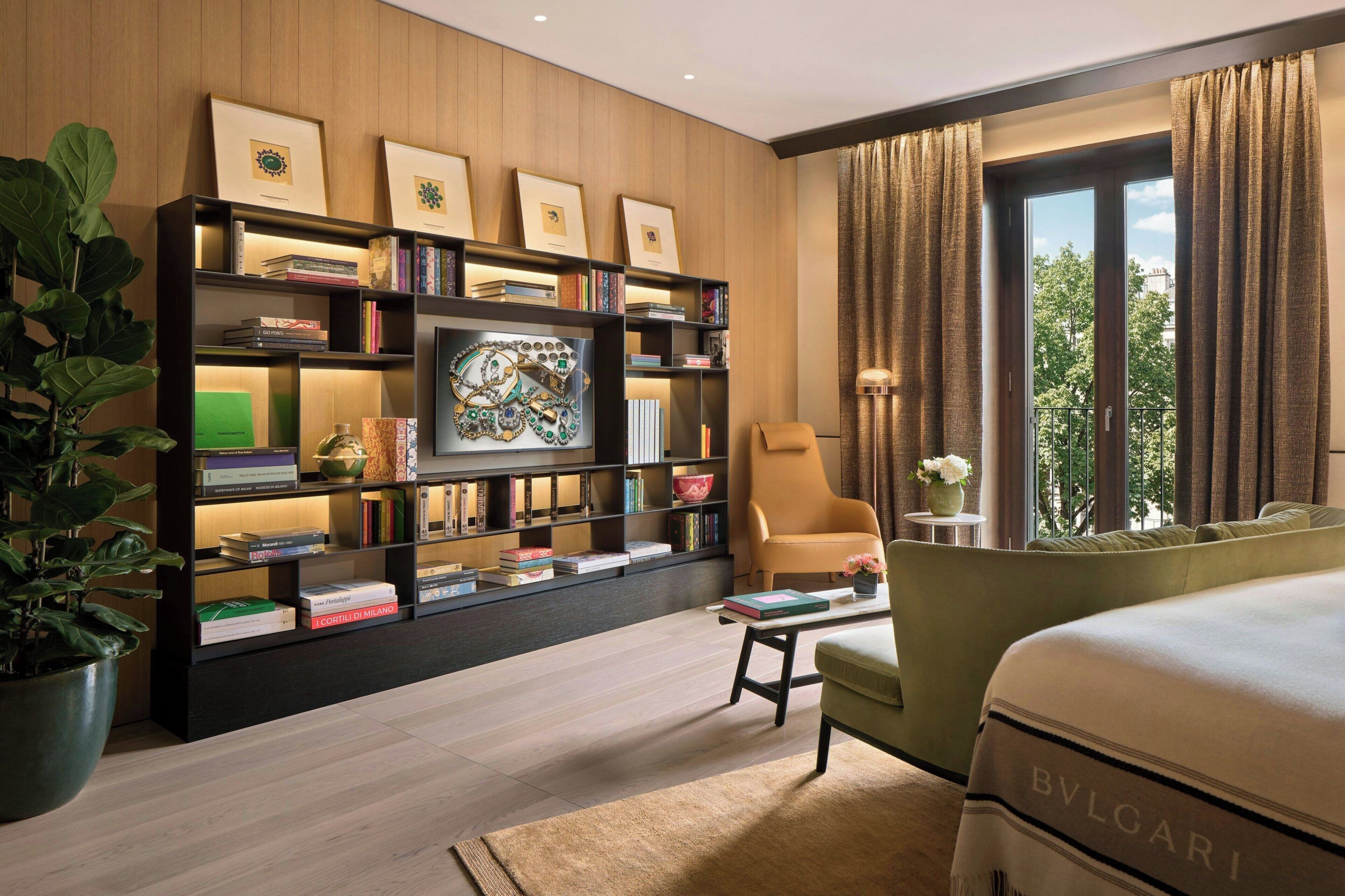 A modern hotel room with a large bed, a sofa, a bookshelf, and a view of the outdoors.