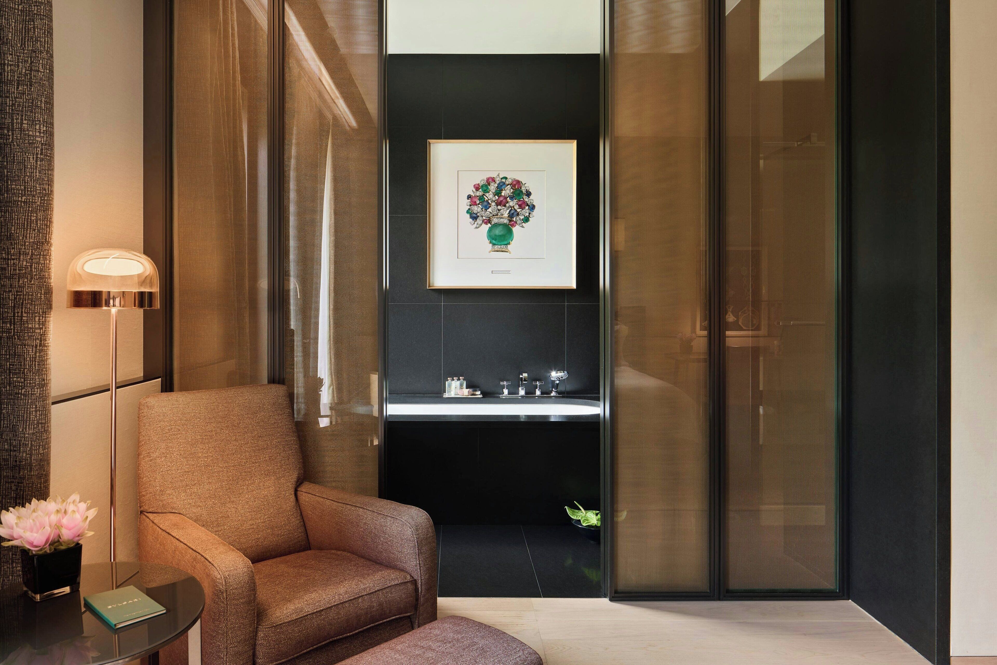 A modern room with a brown armchair, a small table with a book and a vase of flowers, and a framed artwork above a black shelf with a sink.