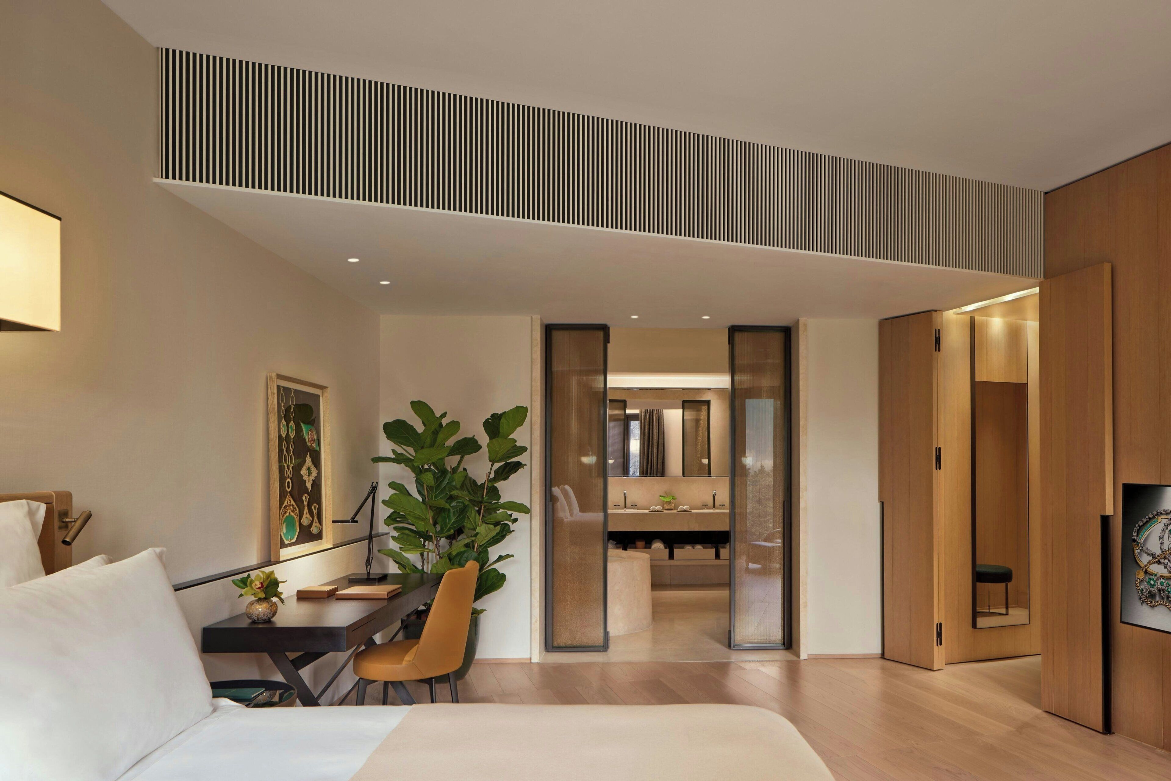 A modern hotel room with a bed, a desk, a chair, a large plant, and a bathroom visible through a glass door.