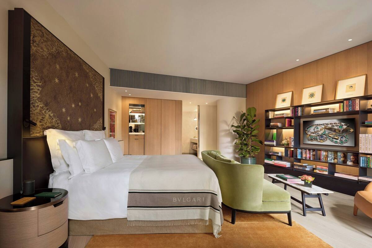 A modern bedroom with a large bed, a green armchair, a bookshelf, and a wall-mounted artwork.
