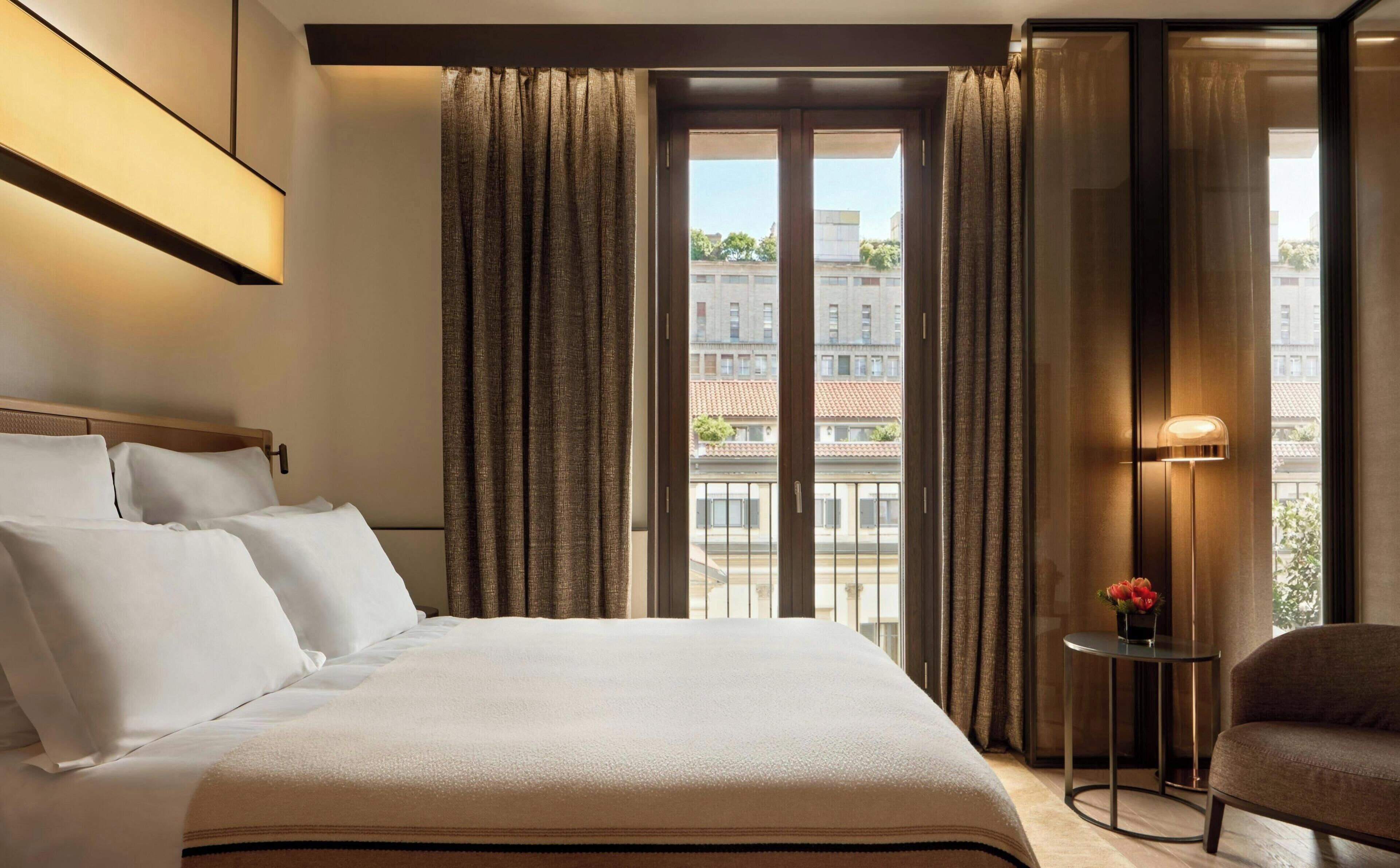 A hotel room with a large bed, a small table with a vase, and a view of buildings through the window.