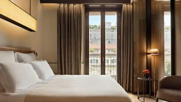 Habitación de hotel con una cama grande, una mesita con un florero y vistas a edificios a través del ventanal.