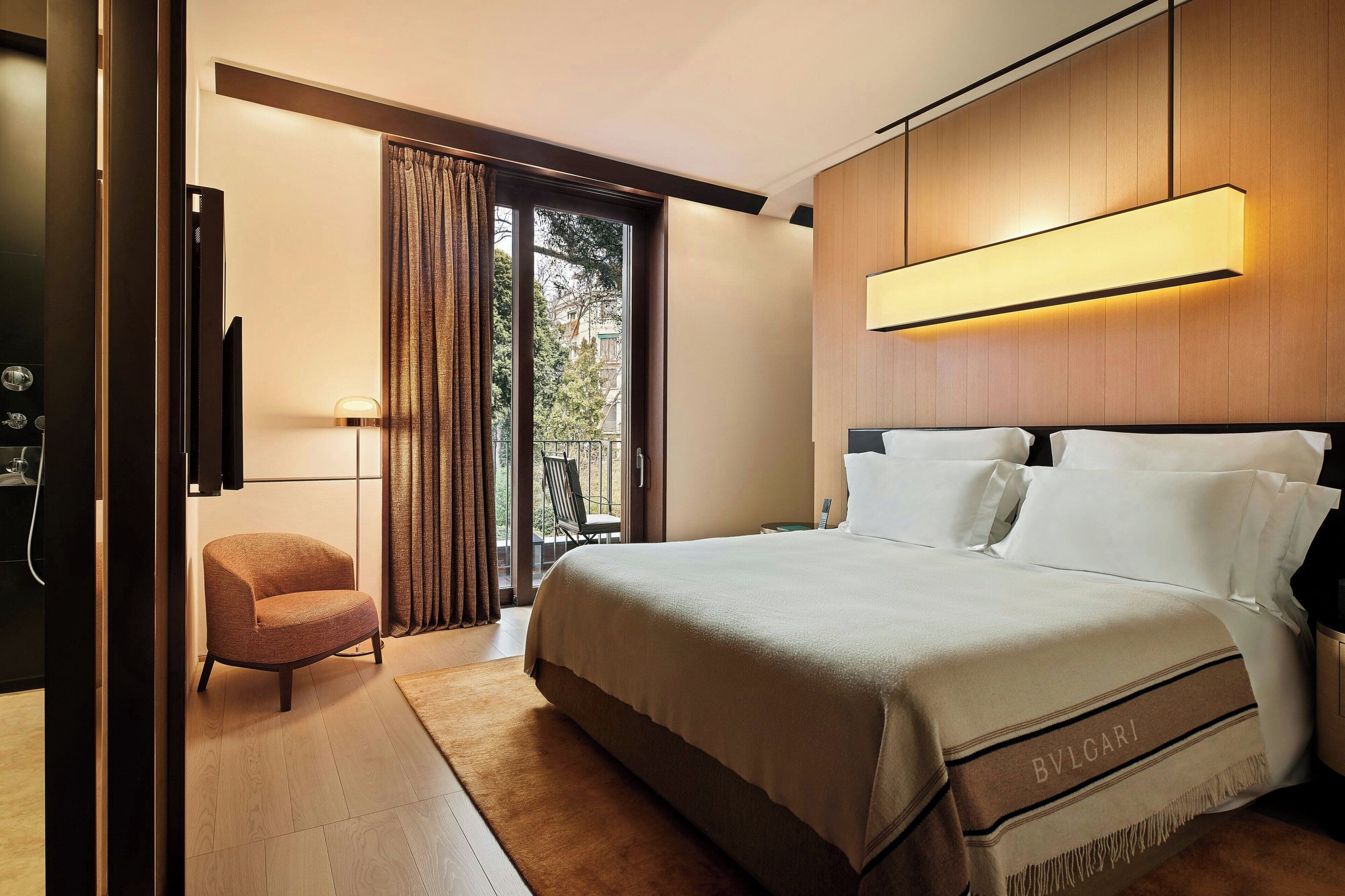 Une chambre d’hôtel moderne dotée d’un grand lit, d’un coin salon avec un fauteuil, d’une télévision et d’un balcon avec des rideaux.