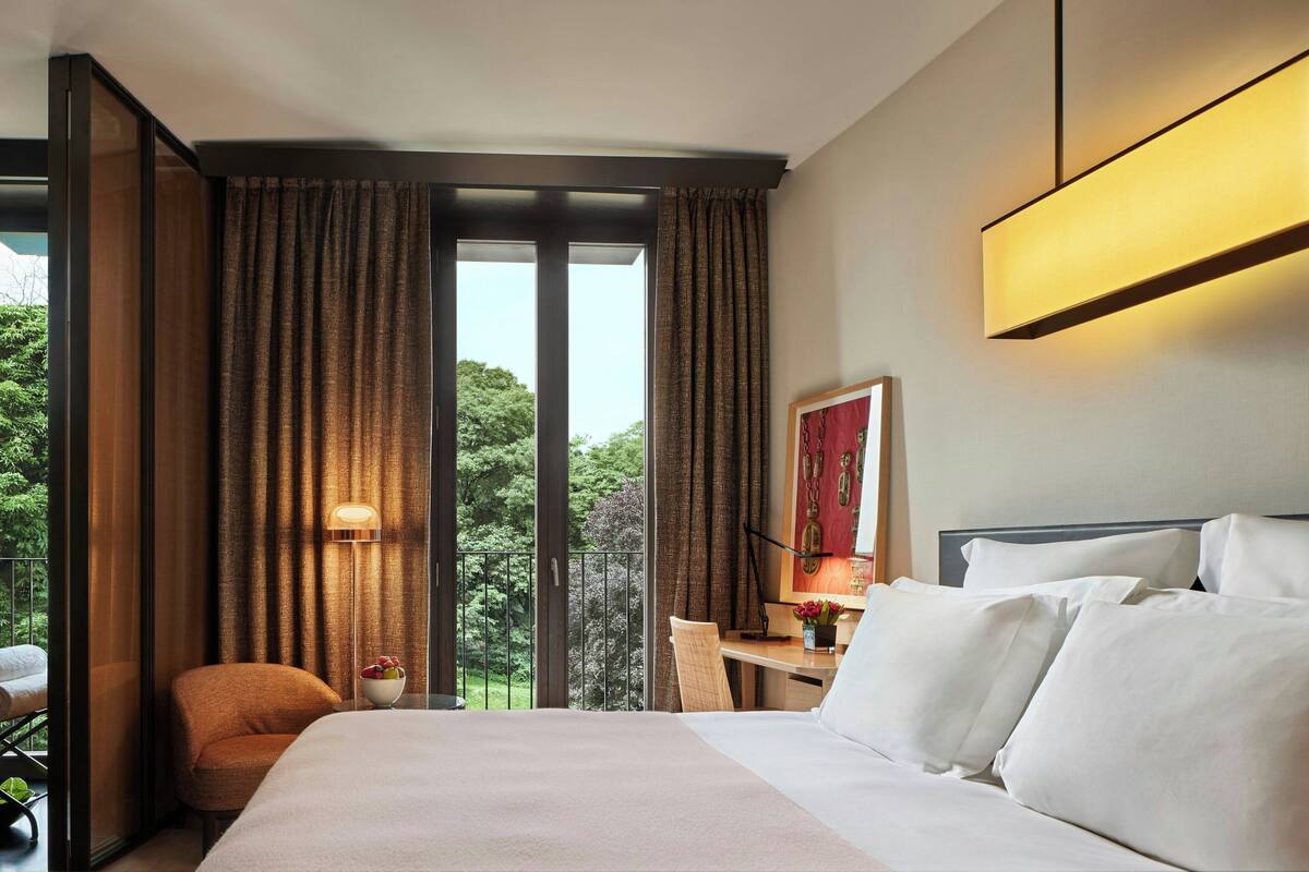 A hotel room with a large bed, a desk with a chair, a window with curtains, and a balcony with a view of trees.