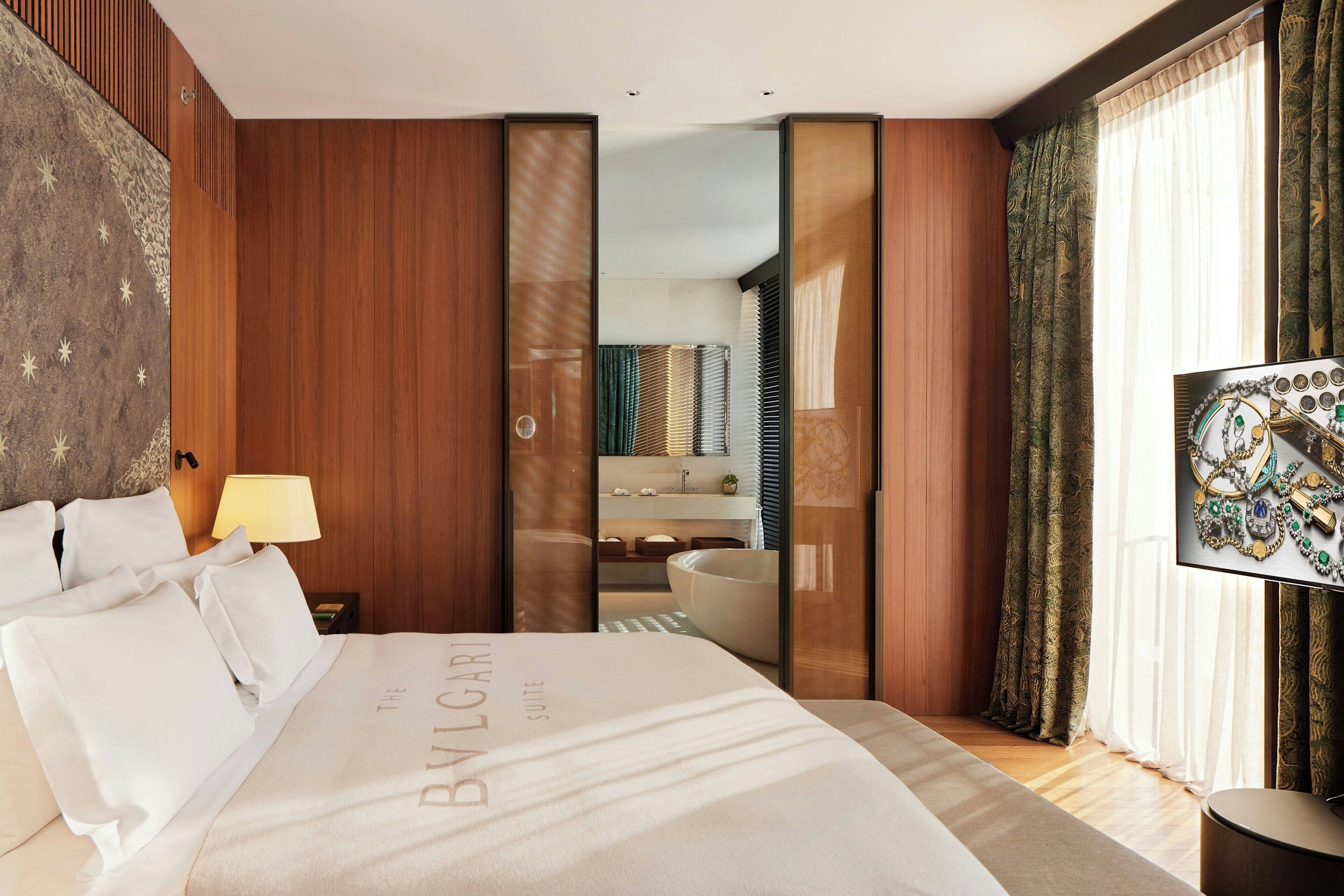 Una habitación de hotel con una cama grande, paneles de madera, un espejo, un baño con bañera y un televisor en la pared.
