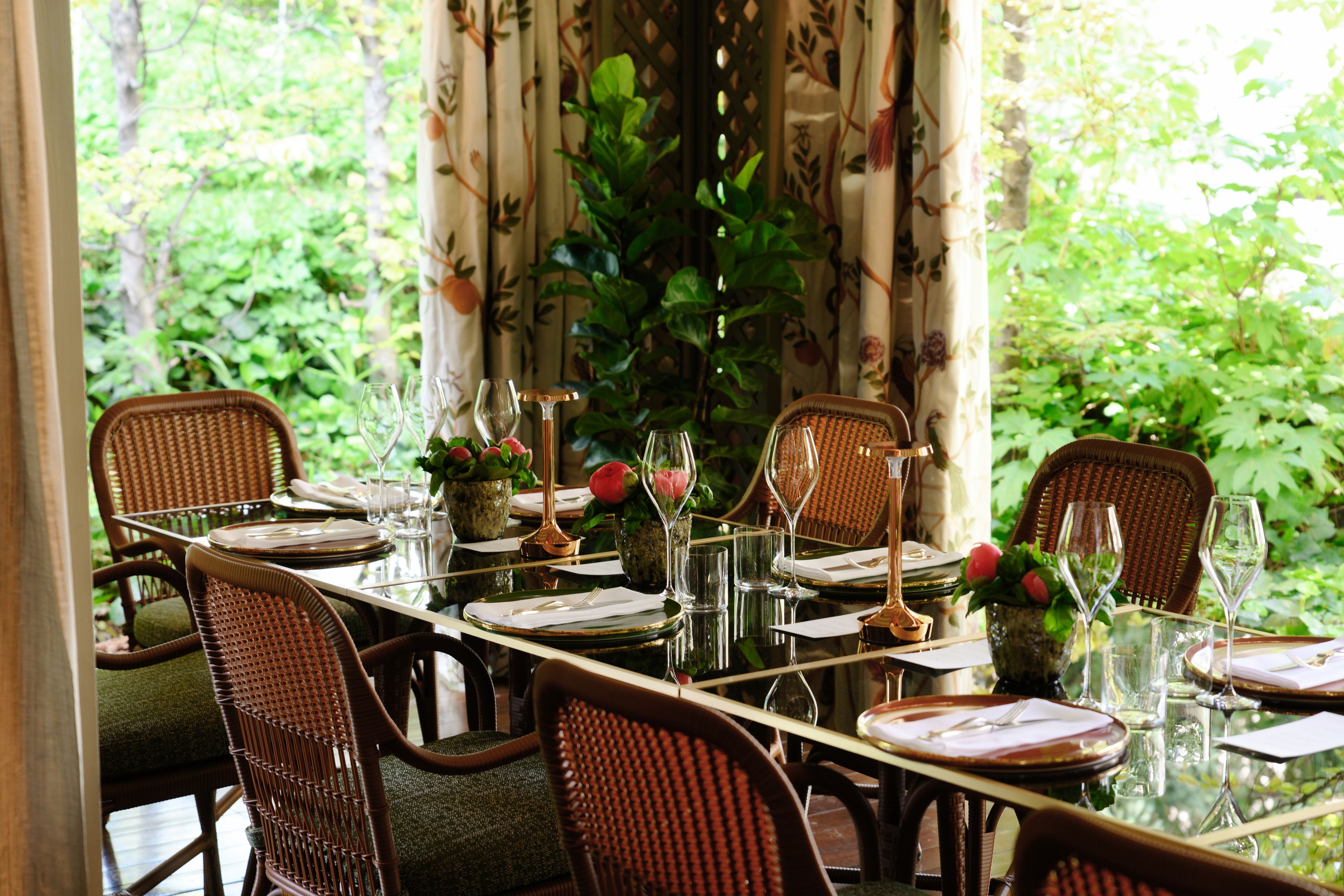 Una mesa de comedor dispuesta con cristalería, platos y cubiertos, rodeada de sillas de mimbre, con vistas a la vegetación a través de cortinas.