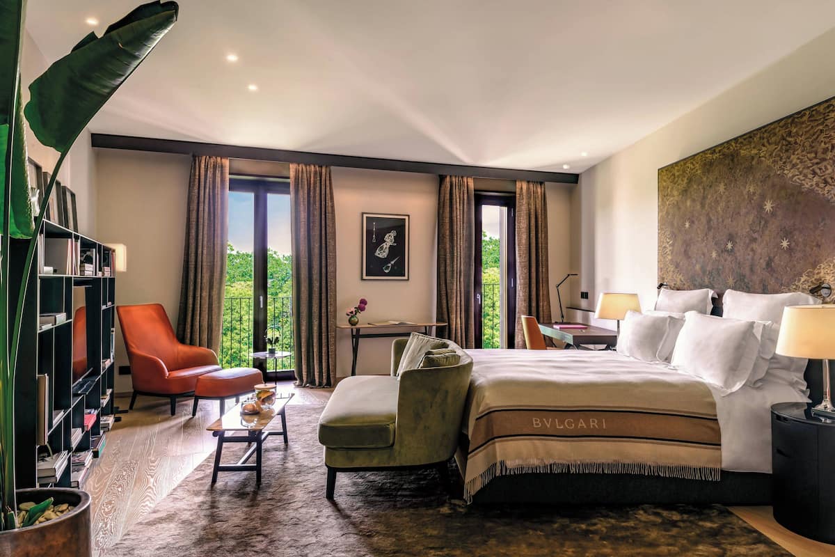 A spacious hotel room with a large bed, a sitting area with a chair and table, a bookshelf, and a view of greenery outside.