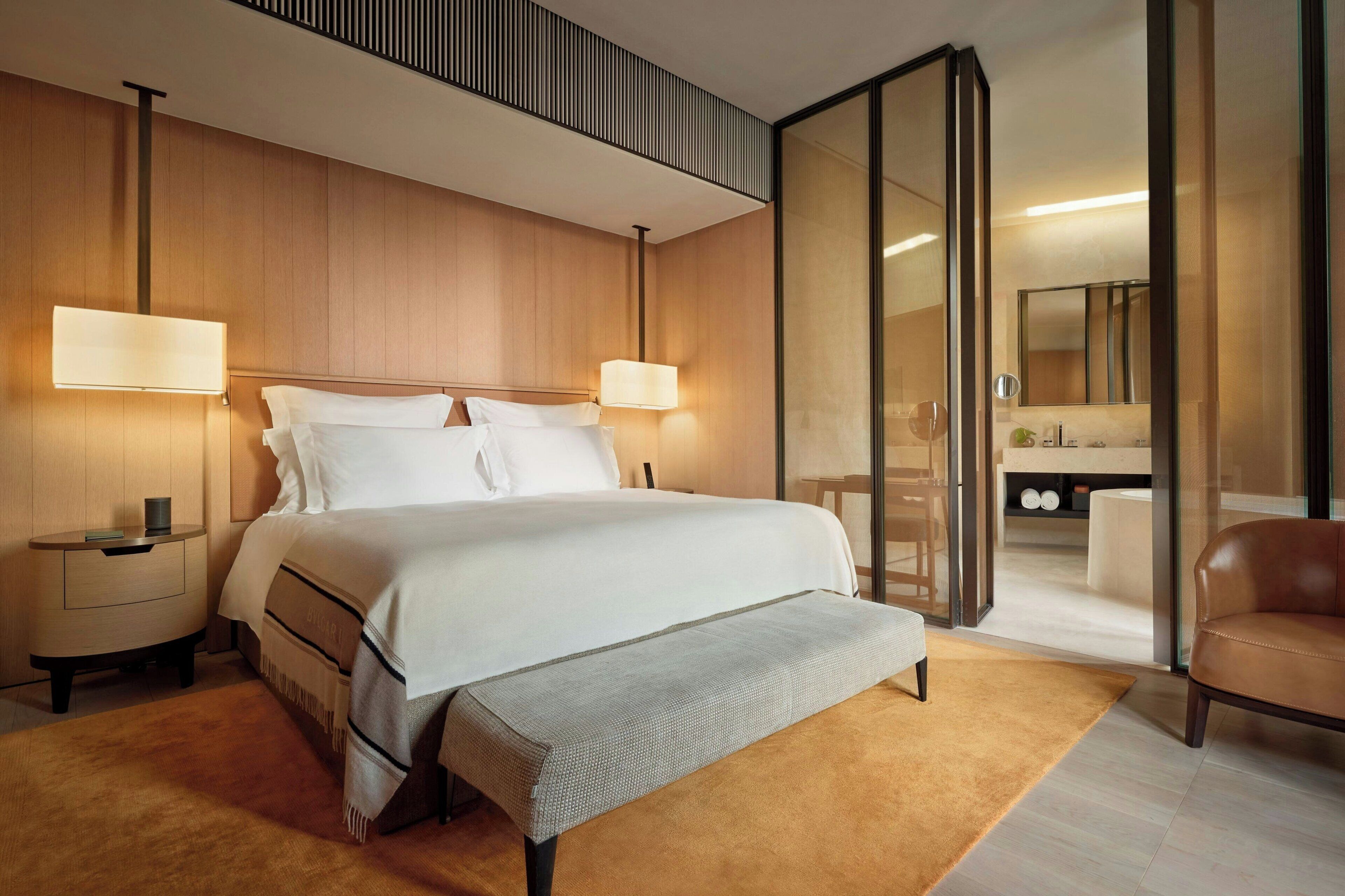 Een moderne hotelkamer met een groot bed, nachtlampjes, een klein nachtkastje, een bankje, een leren fauteuil, en een glazen wand die leidt naar een badkamer.
